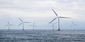 Photograph of an offshore wind farm featuring six wind turbines positioned in the ocean under a cloudy sky.
