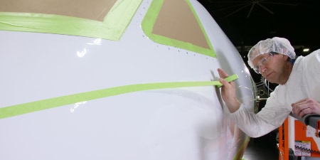 Worker applying masking tape to the nose of an aircraft whose cockpit windows are already masked
