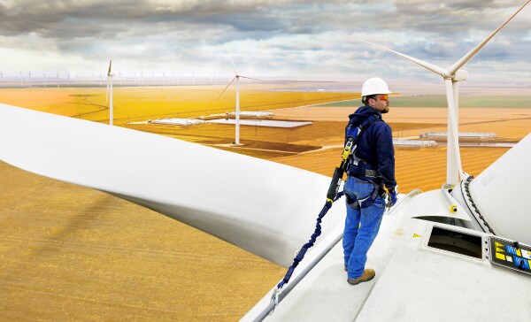 Wind turbine technician wearing industrial safety gear standing on a turbine nacelle overlooking a wind farm.
