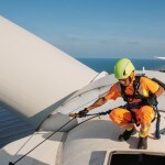 Image of a worker wearing orange safety gear and a yellow helmet inspecting a large white wind turbine blade
