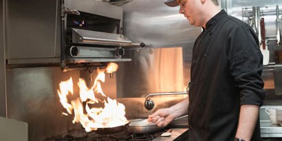 Two restaurant workers cook with a flaming pan and grab food from a bin in a commercial kitchen.
