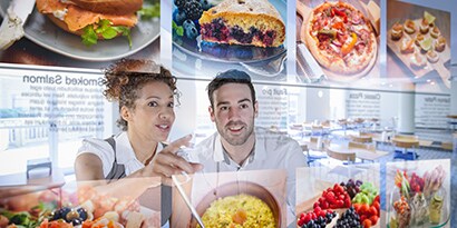 A woman and a man eagerly view floating images of meal choices in a modern restaurant.
