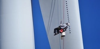Photograph of a worker suspended by ropes performing maintenance on a large white wind turbine against a clear blue sky. 
