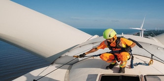 Photograph of a worker in orange safety gear and a yellow helmet inspecting or maintaining a large wind turbine blade near a body of water..

