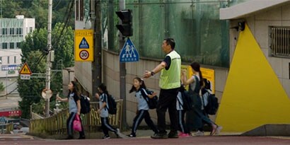 Children cross a street, directed by an adult in a fluorescent safety vest
