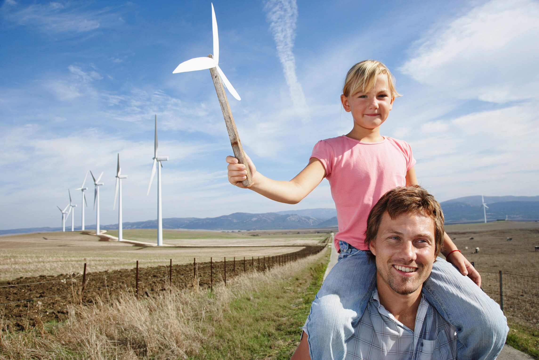 Daughter on father's shoulder holding a pinwheel standing in front of wind farm

