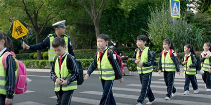 Schoolchildren wearing safety vests cross a crosswalk with adult holding a school zone safety sign
