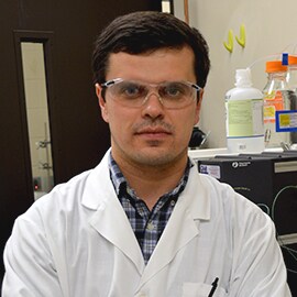 Alexei Voloshin wearing a white lab coat and safety glasses looks directly at camera