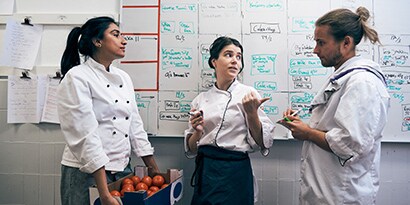 Three restaurant workers chat in front of a whiteboard with complex scheduling information.
